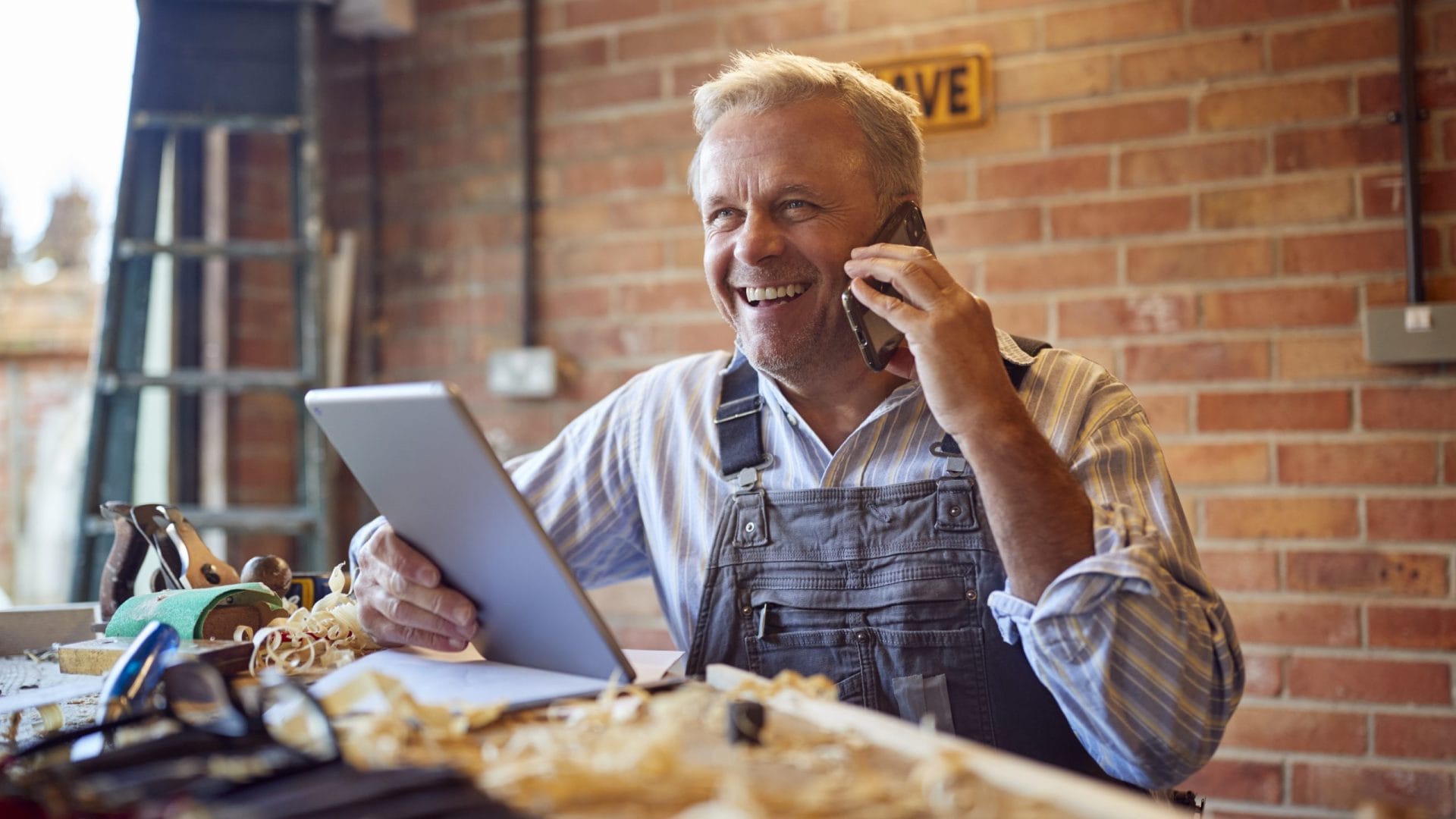 Mature Male Carpenter In Garage Workshop Talking On Mobile Phone