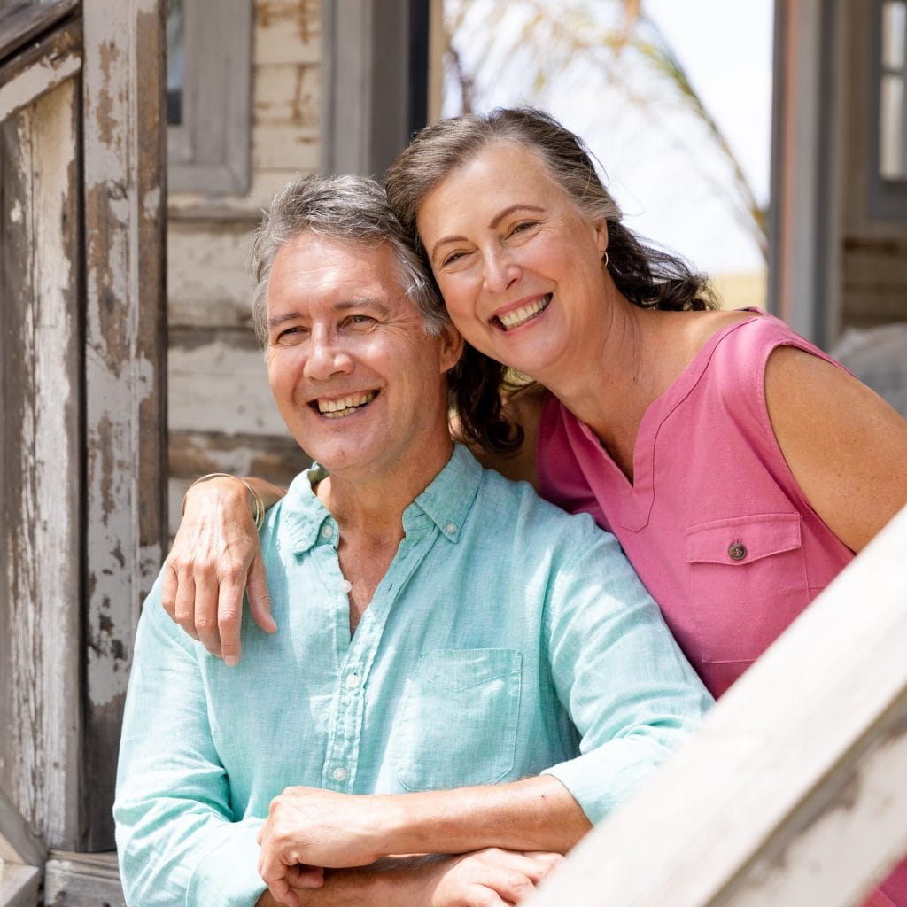Caucasian senior couple smiling cheerfully and sitting on staircase outside cottage. Unaltered, retirement, love, together, vacation, tourist resort, lifestyle, relaxation and enjoyment concept.